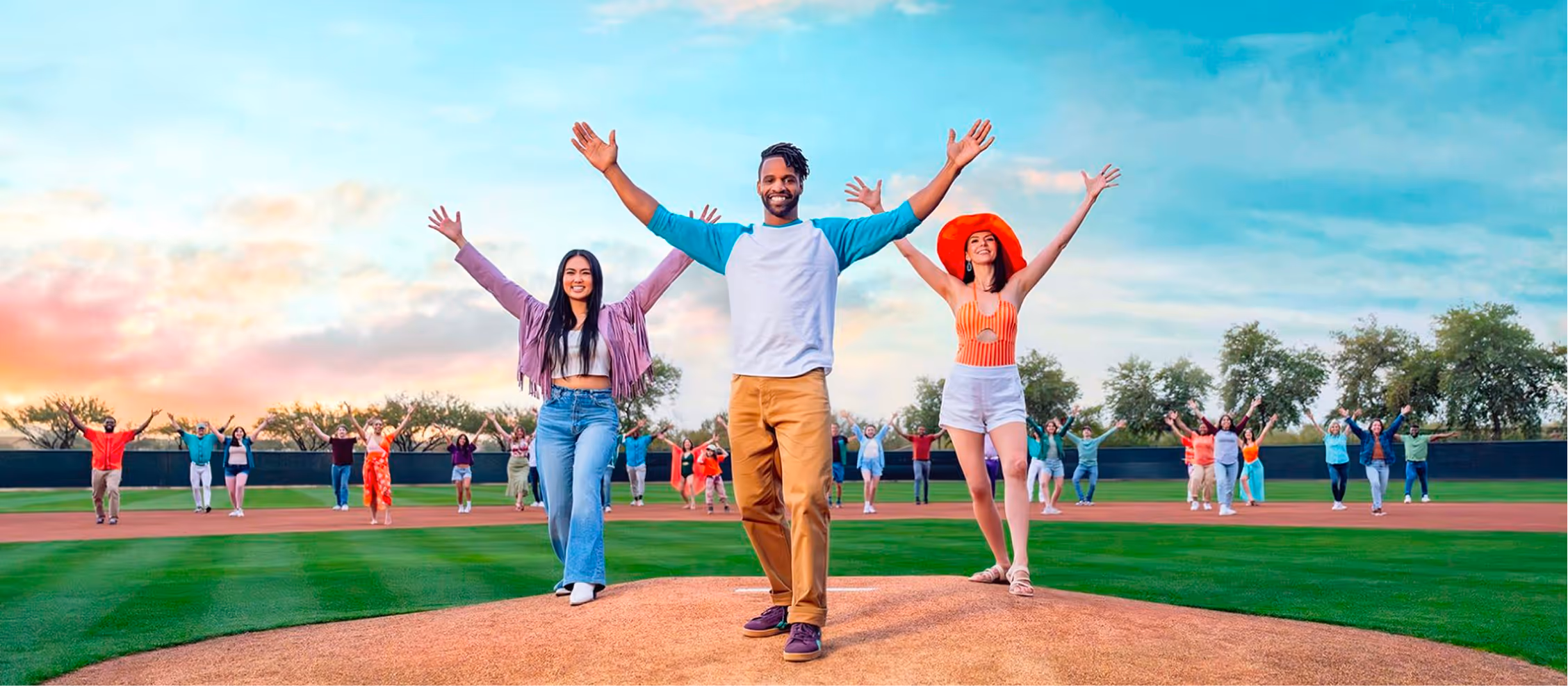 Diverse group on baseball field celebrating with raised arms at sunset